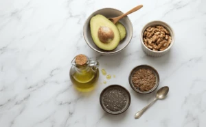 Plant-Based Athlete Nutrition: 7 Proven Strategies to Unlock Elite Performance 3 Healthy fat sources for plant-based athlete nutrition including sliced avocado, walnuts, chia seeds, flaxseeds, and a small jug of cold-pressed olive oil arranged on a marble surface.